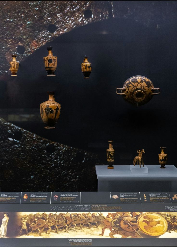 박물관에 전시된 고대 그리스 도자기, 꽃병, 인물은 어두운 배경을 배경으로 전시되어 있으며 아래에는 정보용 명판이 있습니다.