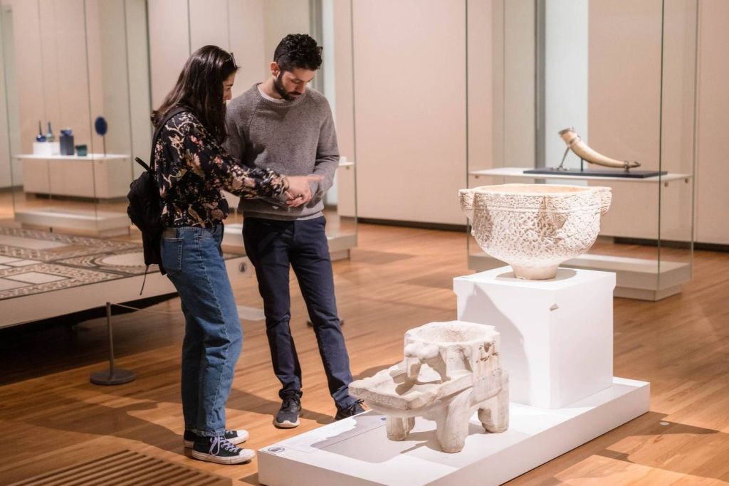 박물관에 있는 두 사람이 스마트폰을 보고 있습니다. 주변에는 다양한 고대의 유물들이 전시되어 있습니다.