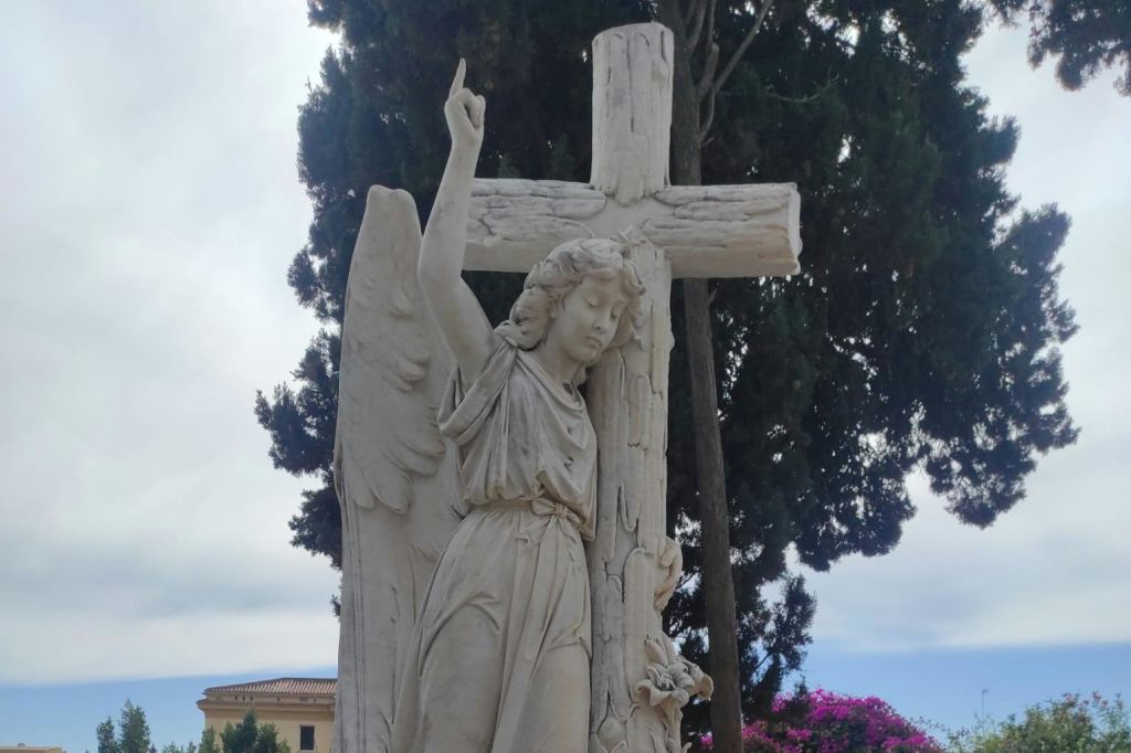 나무와 건물을 배경으로 석조 천사가 십자가 옆에 서 있습니다.