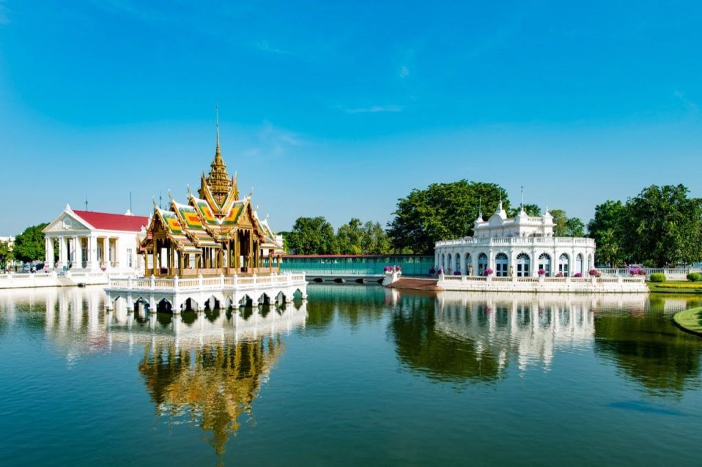 나무를 배경으로 맑고 푸른 하늘 아래 반사 연못이 있는 태국 전통 파빌리온과 흰색 건물입니다.