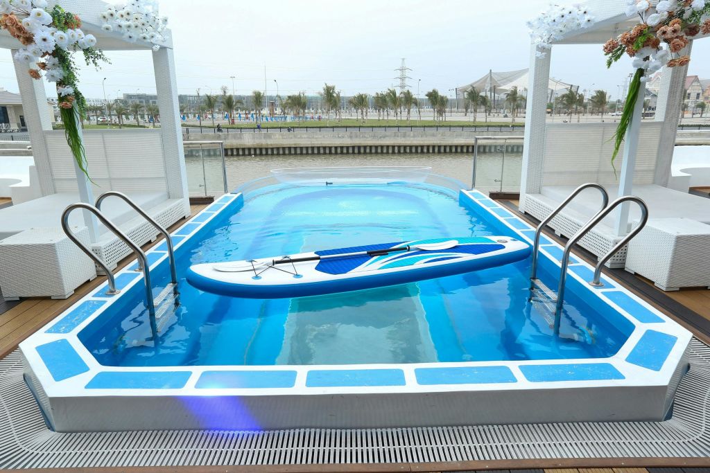 작은 직사각형 수영장에 패들보드가 떠 있으며, 양쪽에 계단이 있습니다. 나무와 구조물이 있는 야외 공간이 배경입니다.