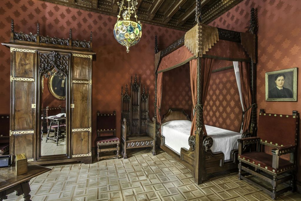 4주식 침대, 등받이가 높은 의자 2개, 나무 바닥, 무늬 벽지, 샹들리에, 액자에 담긴 초상화가 있는 화려한 침실입니다.