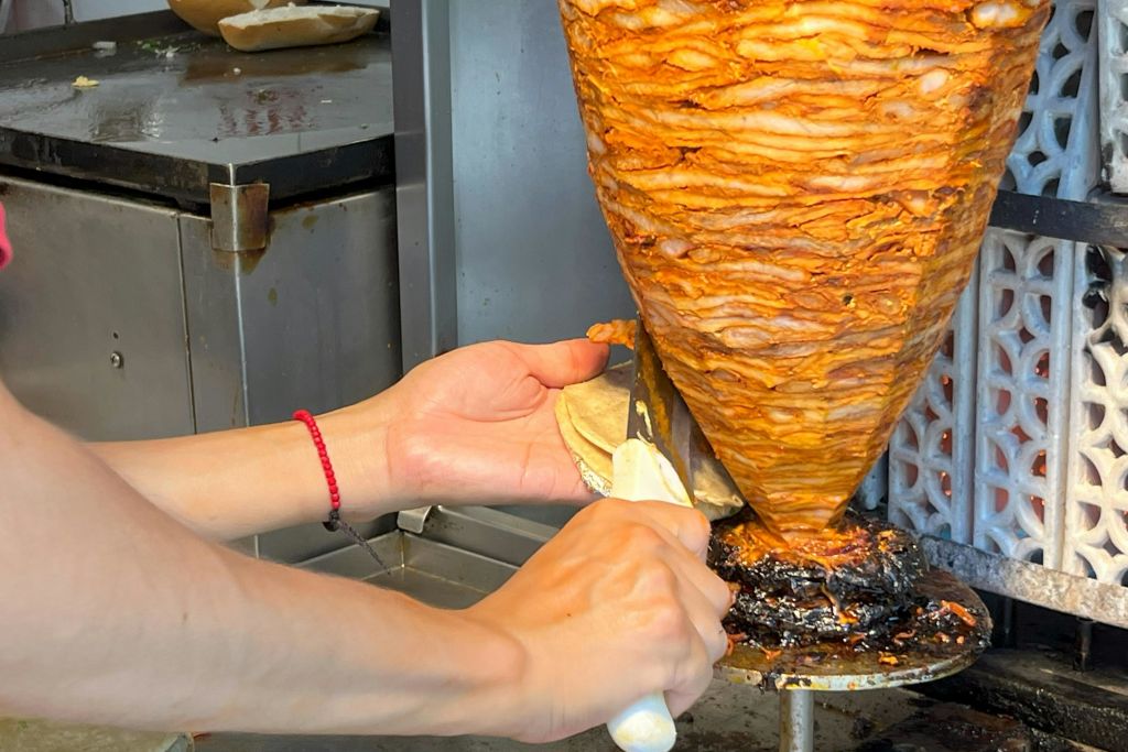 타케로는 양념한 돼지고기를 수직 침에서 토르티야로 썰어 전통 타케리아에서 타코 알 파스토르를 준비합니다.