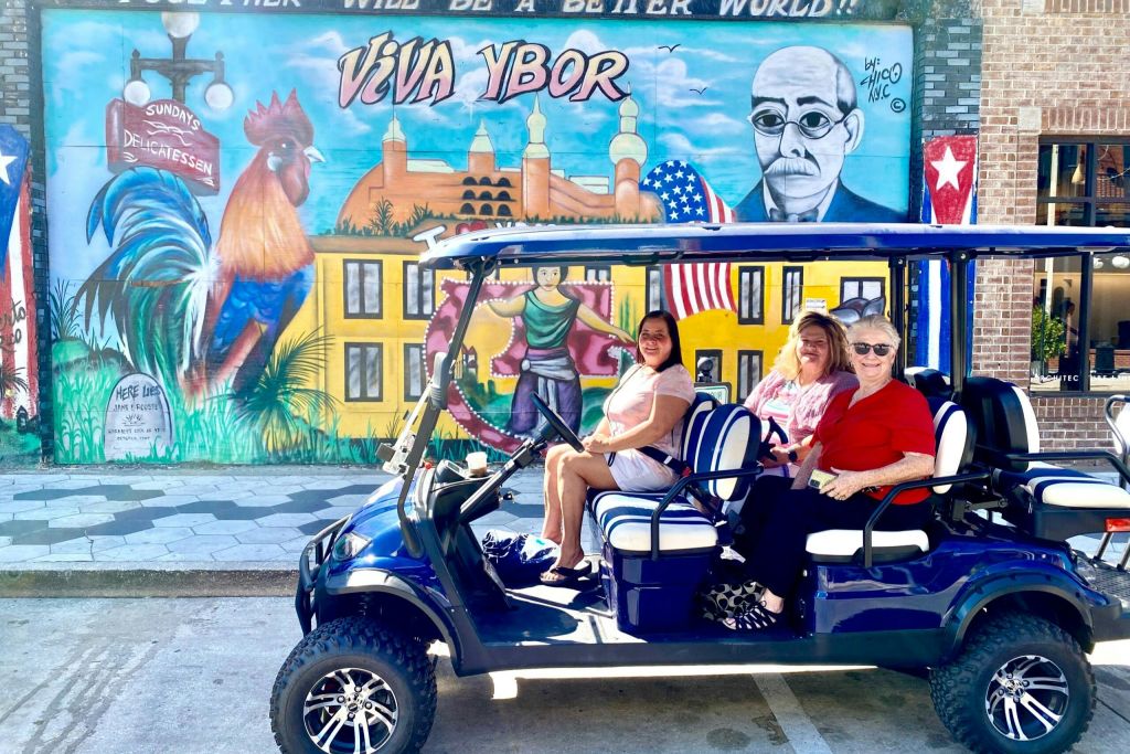 건물, 깃발, 벽돌 벽에 그려진 초상화가 있는 화려한 벽화 앞에 세 명의 여성이 파란색 골프 카트에 앉아 있습니다.