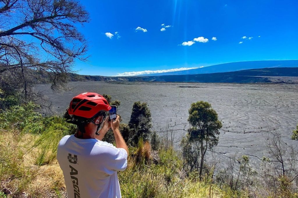 빨간 헬멧을 쓴 사람이 나무가 흩어져 있는 광활하고 바위가 많은 풍경과 위쪽의 밝고 푸른 하늘을 사진으로 찍고 있습니다.