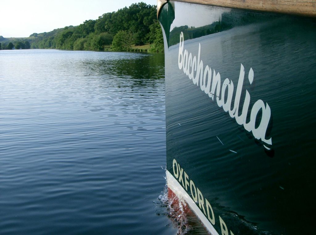 "옥스포드" 가 보이는 "바카날리아" 라는 이름의 녹색 보트의 측면은 숲이 우거진 해안가 근처의 잔잔한 물을 통과합니다.