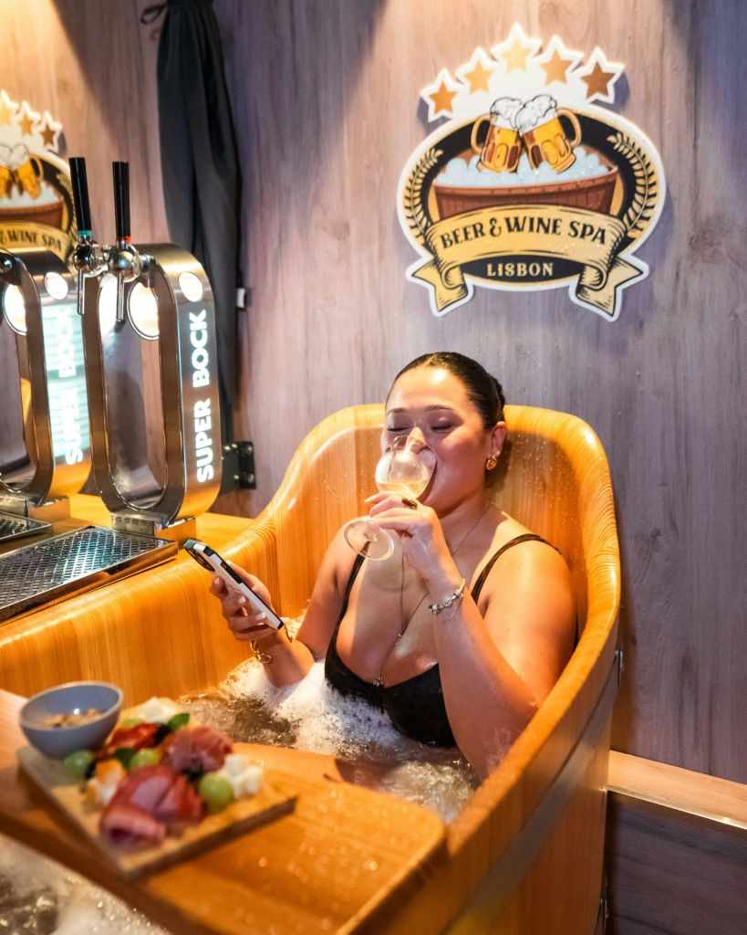 한 여성이 맥주와 와인 스파에서 나무 욕조에서 휴식을 취하며 와인을 마시고 휴대폰을 들고 근처에 음식과 맥주 탭이 있습니다.