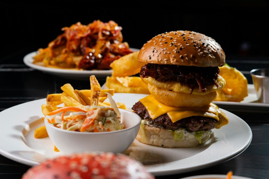 참깨 롤빵에 양파 링과 바베큐 소스를 곁들인 치즈버거로 흰 접시에 감자튀김과 코울슬로를 곁들입니다.