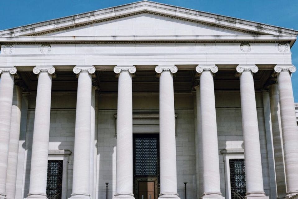 6개의 커다란 기둥이 페디먼트를 받치고 있는 신고전주의 양식의 건물 외관으로, 문에 화려한 그릴이 있는 메인 출입구가 있습니다.