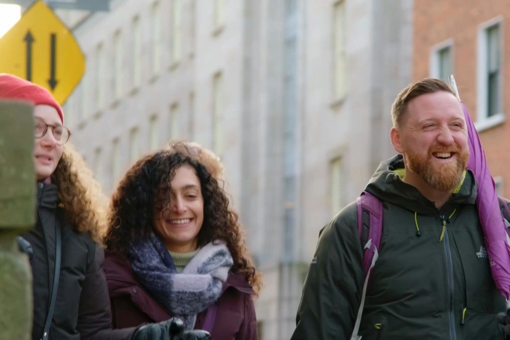 재킷과 배낭을 두른 미소 짓는 남녀가 높은 건물을 배경으로 도심 속 야외를 거닐고 있습니다.