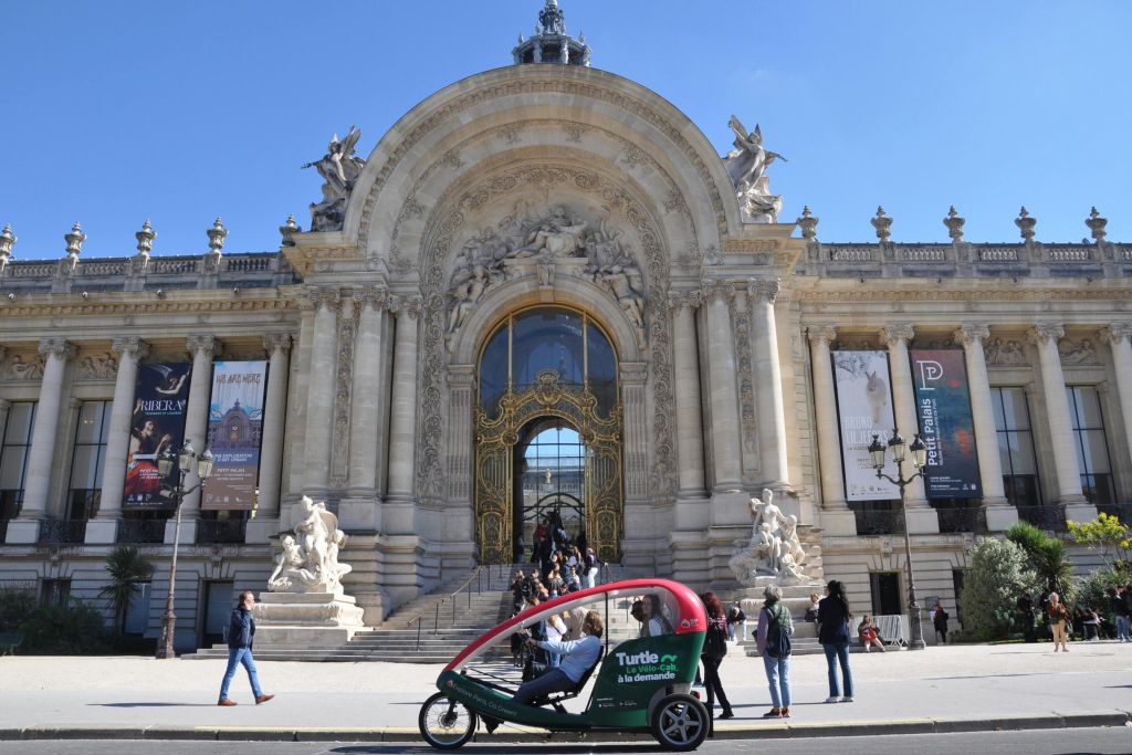 사람들은 걷고 입구에 동상과 대형 배너가 있는 웅장한 건물의 화려한 외관을 지나 빨간색-녹색 페디캡을 타고 지나갑니다.