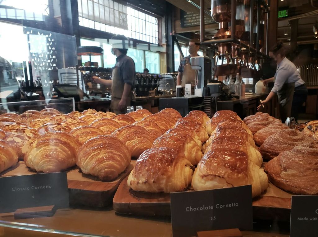 베이커리 진열 케이스에 크루아상과 초콜릿 코르네토가 줄지어 있고, 바리스타가 배경으로 작업합니다.