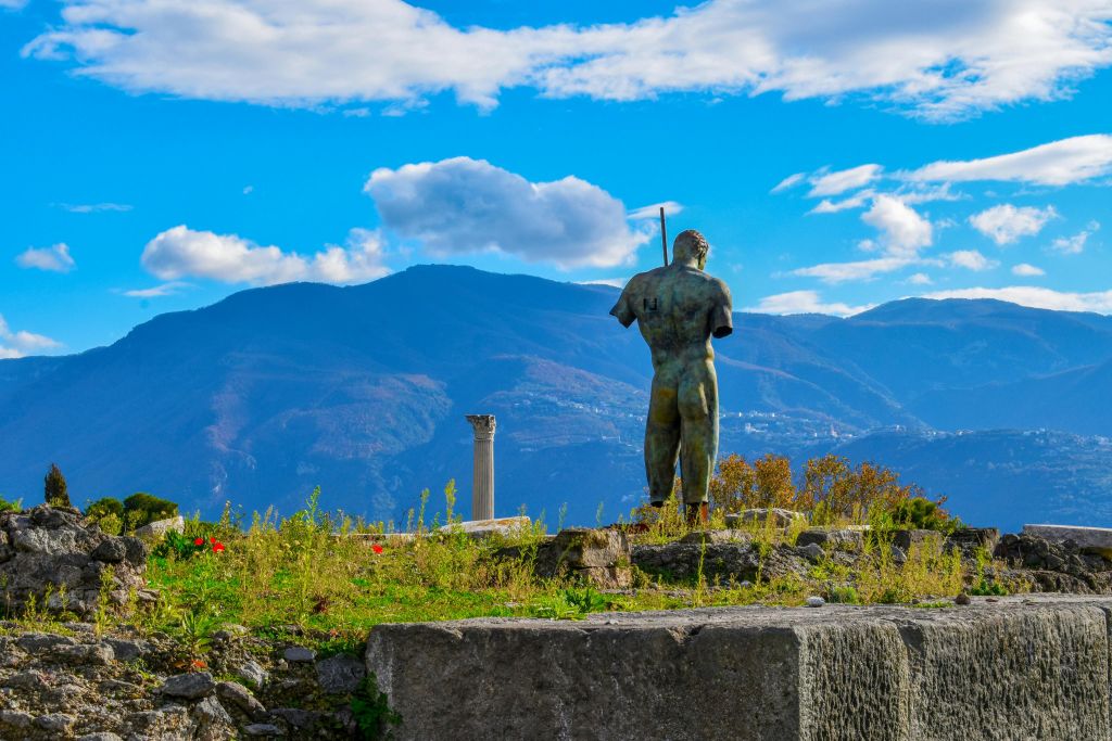 저 멀리 보이는 동상이 고대 유적지에 서 있습니다. 산과 한 개의 기둥이 흩어진 구름과 함께 밝고 푸른 하늘 아래 보입니다.
