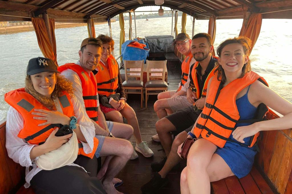 주황색 구명조끼를 입은 세 사람이 보트에 앉아 카메라를 보며 미소를 짓고 있습니다. 배경에는 의자 2개와 지붕이 있는 공간이 있습니다.