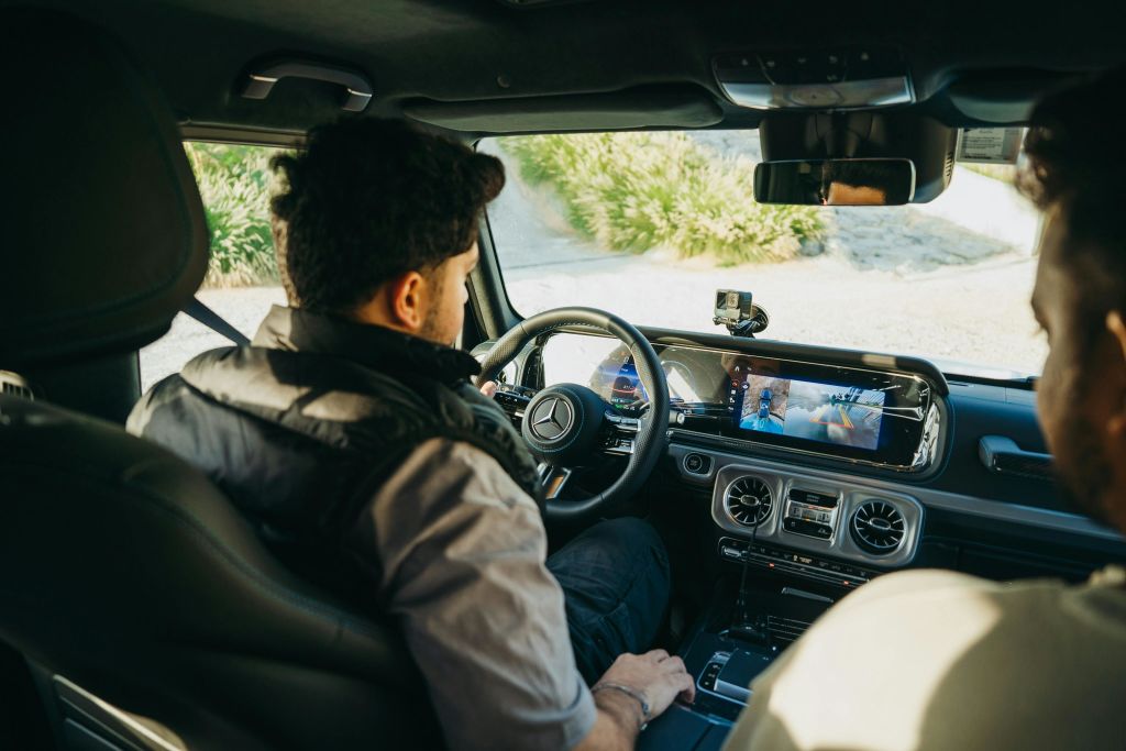 스티어링 휠에 메르세데스-벤츠 로고가 새겨진 차량을 운전하는 회색 셔츠와 검은색 조끼를 입은 남성.