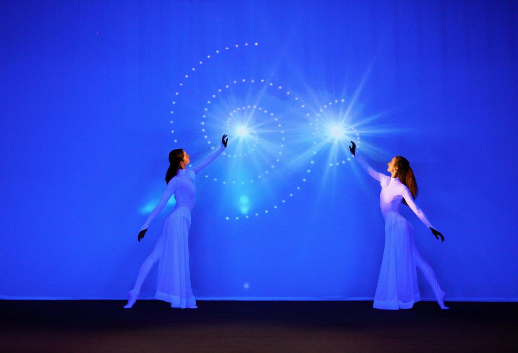 흰색 의상을 입은 두 명의 공연자가 파란색 배경에 빛나는 나선형 빛 패턴을 향해 손을 내밀고 있습니다.