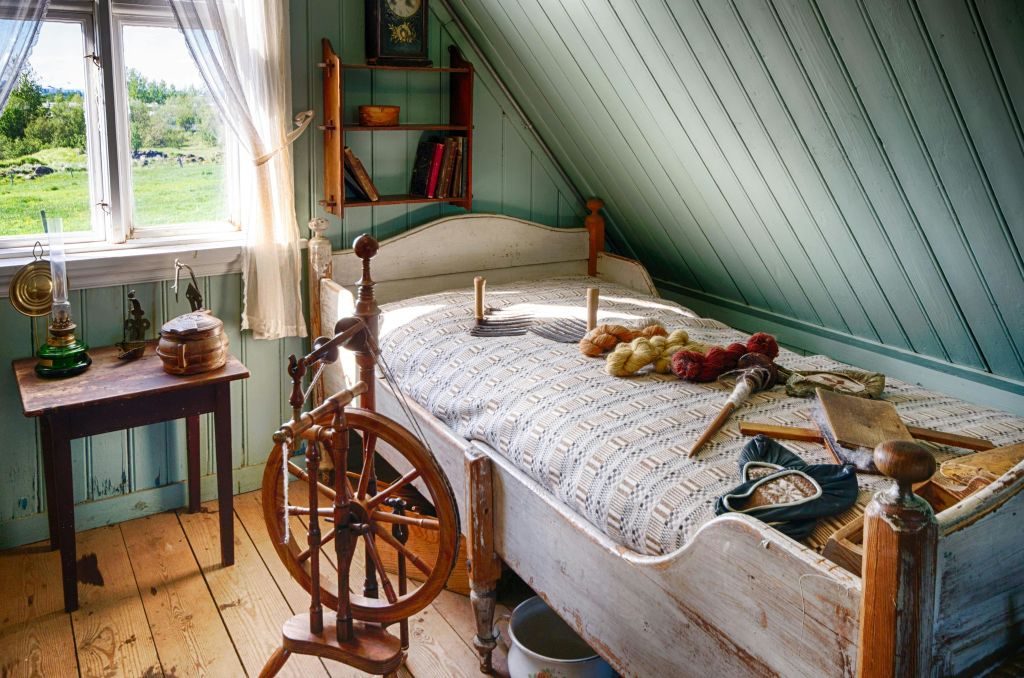 회전 바퀴가 달린 소박한 침실, 뜨개질 담요로 덮인 침대, 원사, 창문 옆 벽 선반에 놓인 책.
