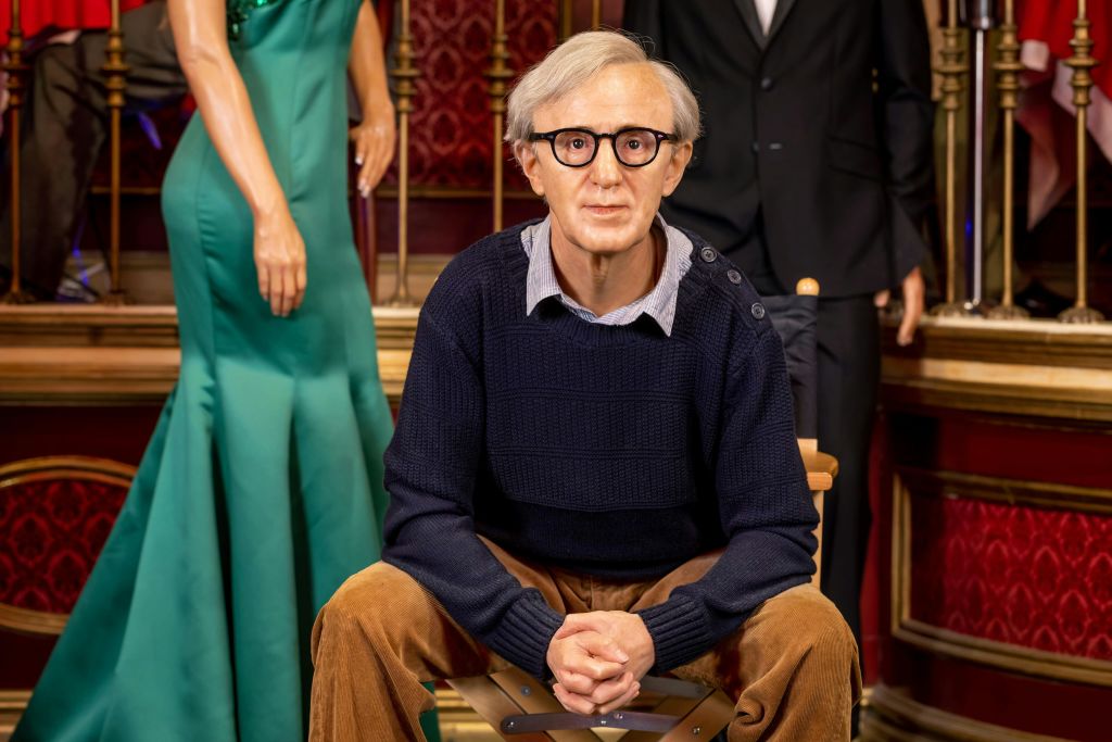 정장 차림의 다른 왁스 피규어에 둘러싸인, 안경과 스웨터를 착용한 노인의 왁스 피규어.