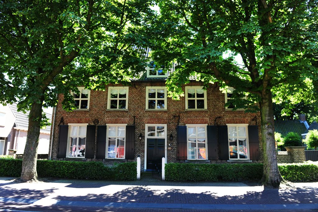 여러 개의 창문, 검은 셔터, 나무가 있는 유서 깊은 벽돌 건물로 앞 보도를 따라 그늘이 있습니다.