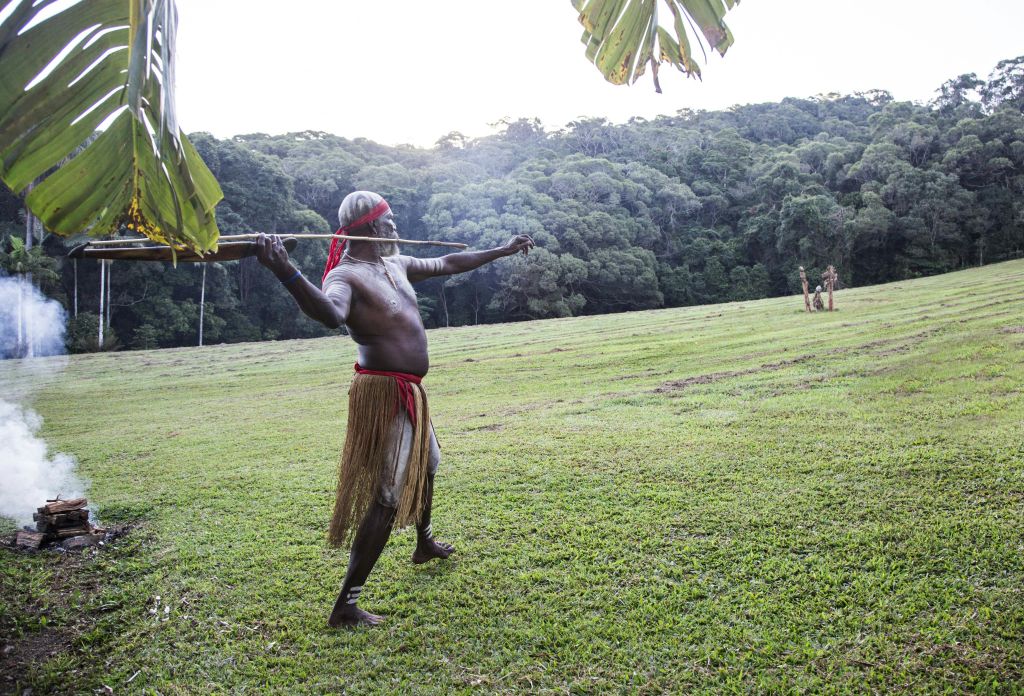 숲이 우거진 언덕을 배경으로 거리를 겨냥한 창을 들고 풀밭에 전통 의상을 입은 남성이 서 있습니다.