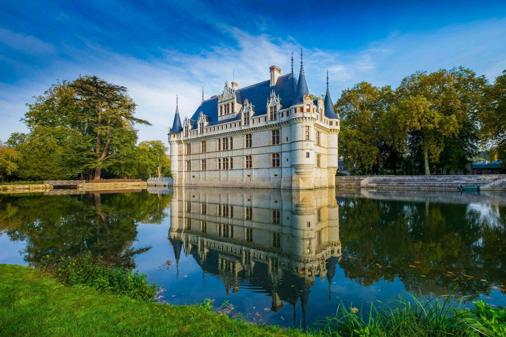 푸른 하늘 아래 나무를 배경으로 여러 개의 뾰족한 탑이 있는 웅장한 성이 잔잔하게 둘러싸인 해자에 반사되어 있습니다.