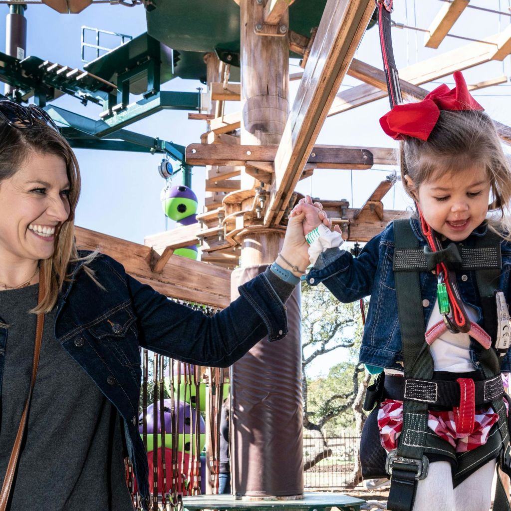 한 여성이 하네스를 착용한 어린 소녀가 웃으며 로프 코스를 탐색하는 것을 돕고 있습니다. 소녀는 머리카락에 커다란 붉은 활을 가지고 있습니다.