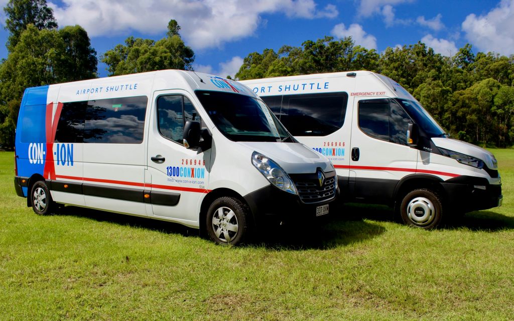 "100% ConXion" 브랜딩이 있는 두 대의 흰색 셔틀 밴이 부분적으로 흐린 하늘 아래 잔디에 주차되어 있습니다.