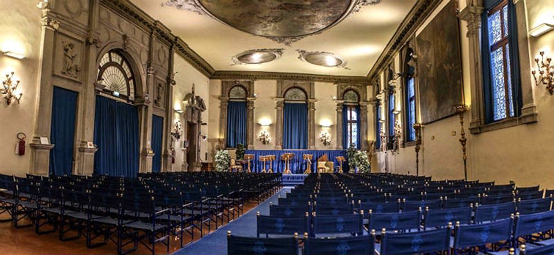 파란색 커튼, 높은 창문, 장식용 식물로 장식된 무대를 마주하고 있는 검은색 의자가 줄지어 있는 화려한 객실입니다.