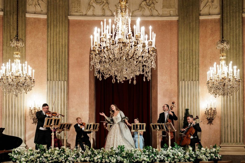 샹들리에가 있는 화려한 홀에서 열리는 클래식 음악 공연. 음악가들은 현악기를 연주합니다. 가수는 의상을 입고 있습니다.