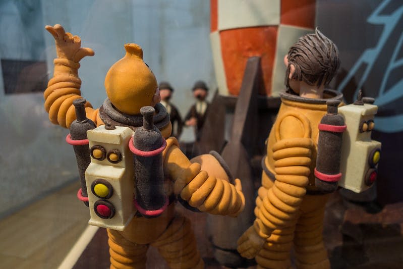 제트팩이 달린 주황색 우주복을 입은 두 개의 인형이 모형 로켓 근처에 서 있습니다. 배경에는 한 무리의 작은 인물들이 보입니다.