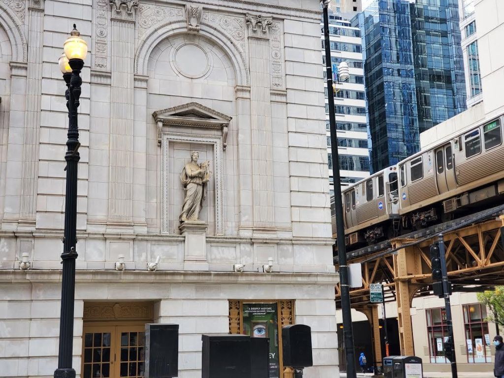 동상, 가로등, 고가 열차가 지나가는 신고전주의 건물 외관이 있는 도시 풍경.