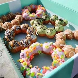 다양한 생동감 넘치는 토핑과 글레이즈가 담긴 모듬 폰 데 링 도넛 상자가 햇빛 아래 야외에 놓여 있습니다.