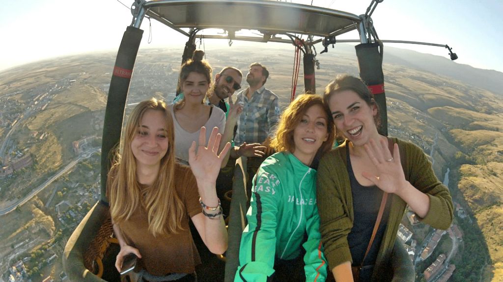 배경에 풍경이 보이는 열기구 바구니 안에서 다섯 명이 미소를 지으며 손을 흔들고 있습니다.