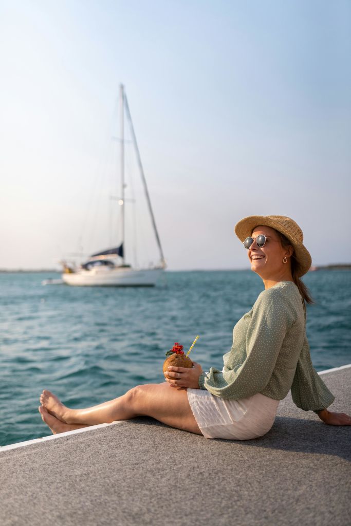 선글라스와 모자를 쓴 여성이 열대 음료를 들고 바다에 앉아 있습니다.