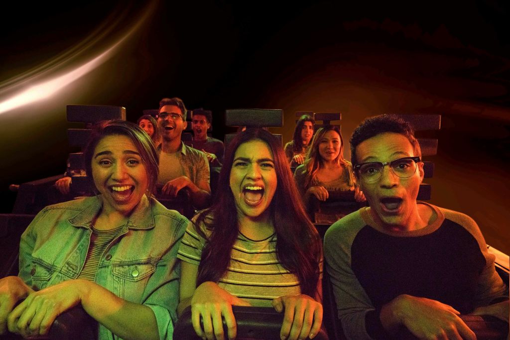 공간을 테마로 한 실내 놀이기구에 앉아 신나게 웃고 비명을 지르는 사람들. 반지가 빛나는 어두운 배경.