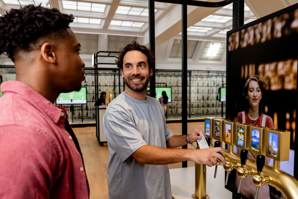 두 남자가 실내에 있습니다. 하나는 터치스크린 키오스크를 작동합니다. 방은 커다란 창문과 사람들이 배경으로 보입니다.