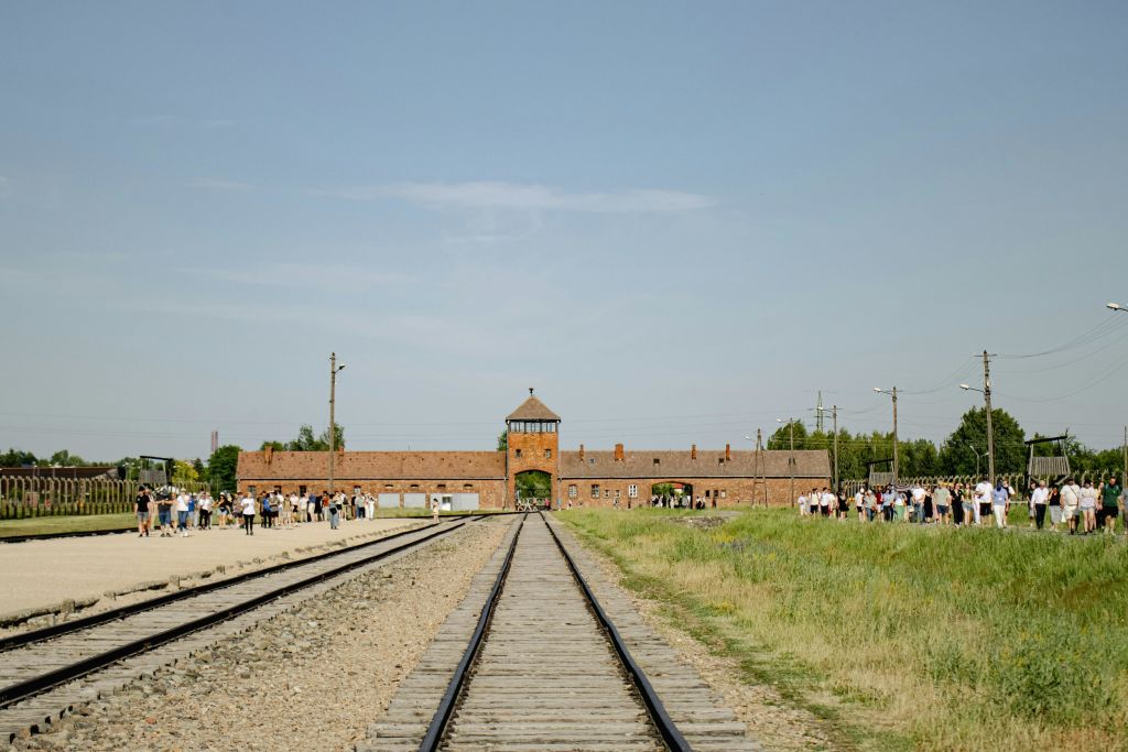 양쪽에 사람들에 둘러싸인 타워가 있는 먼 벽돌 건물로 이어지는 직선 철로. 위쪽의 맑은 하늘.