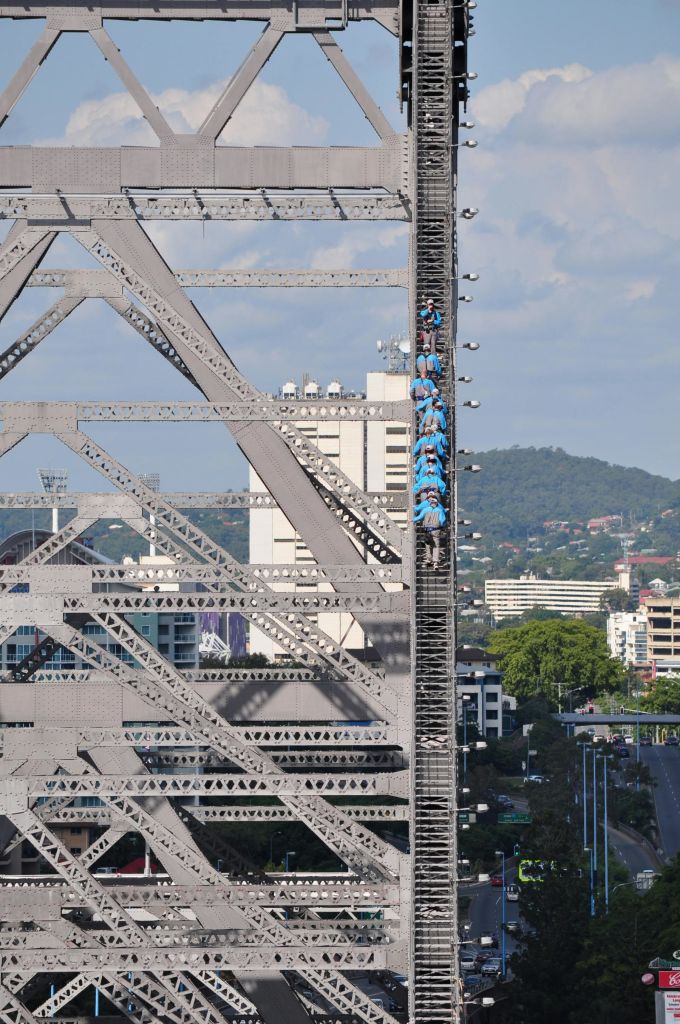 도시 풍경과 언덕을 배경으로 금속 교량 구조물을 오르고 있는 파란색 셔츠를 입은 한 무리의 사람들.