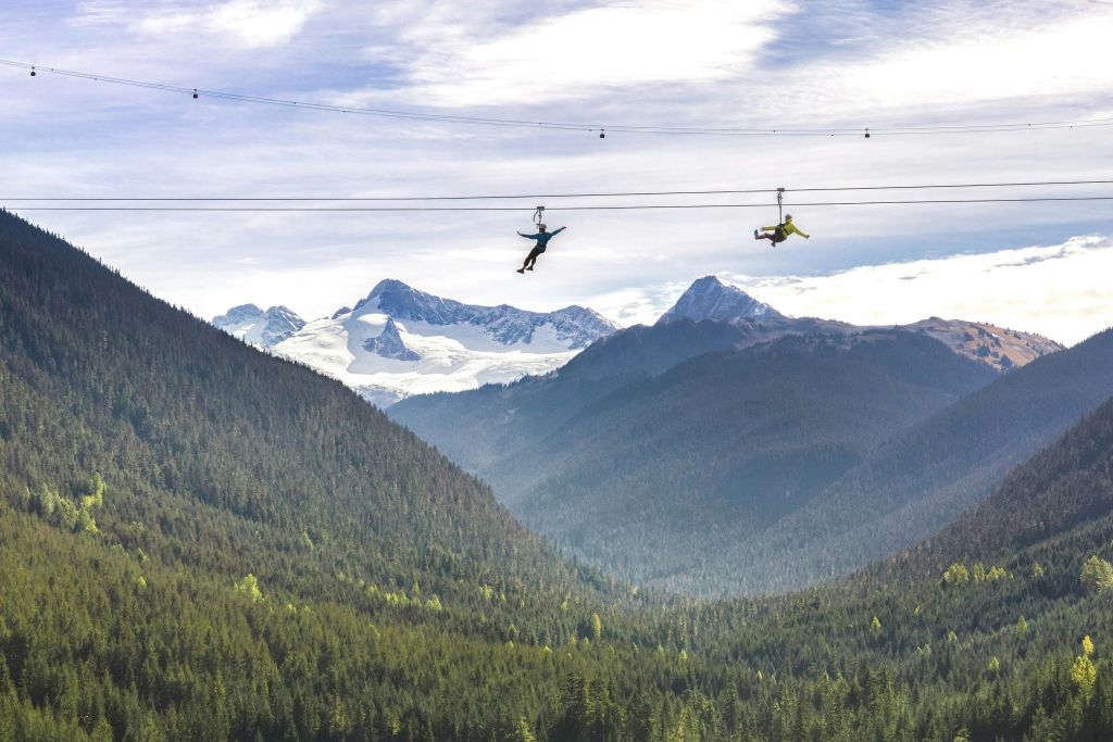 부분적으로 흐린 하늘 아래 눈 덮인 산을 배경으로 울창한 숲을 가로지르는 두 사람.