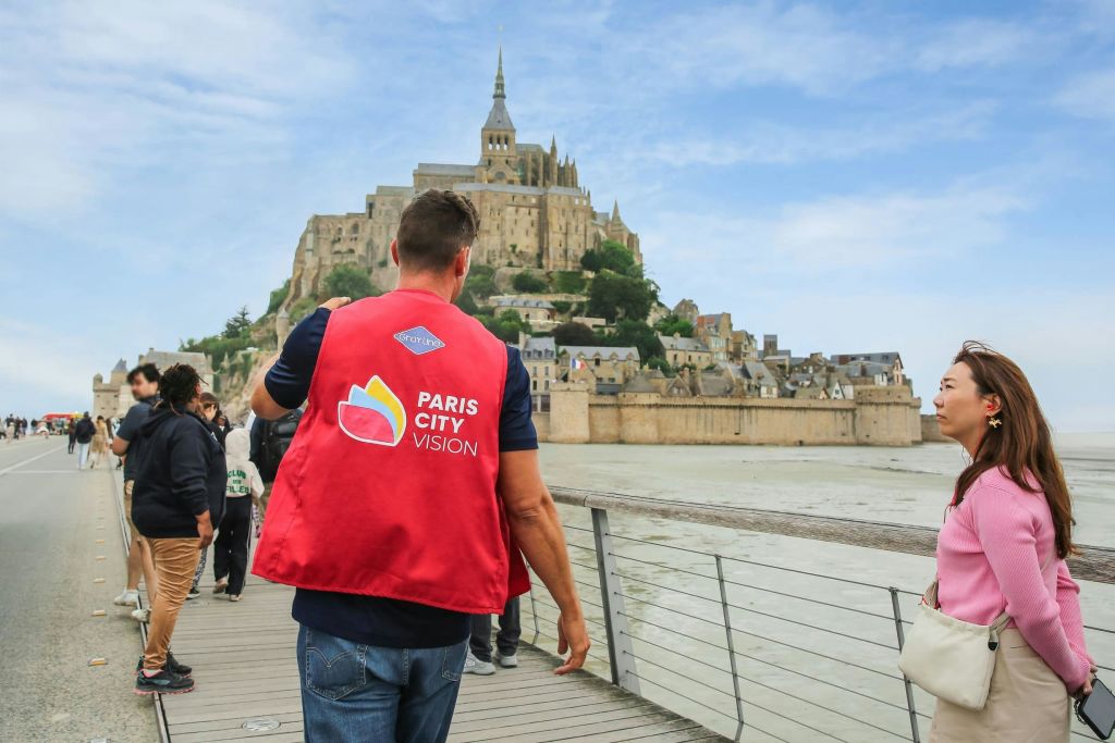 몽생미셸 (Mont Saint-Michel) 이 내려다보이는 다리 위에 두 사람이 서 있습니다. 한 사람은 빨간색 "파리 시티 비전 (Paris City Vision)" 조끼를 입고 있습니다.
