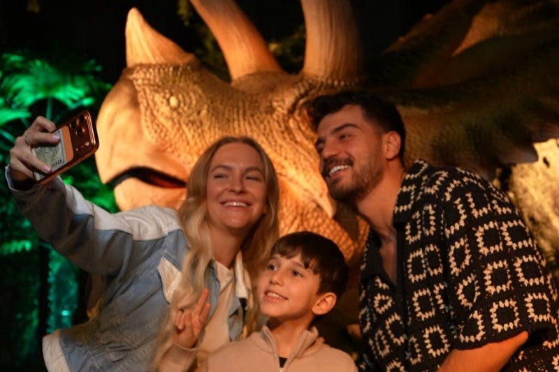 세 사람이 조명이 어두운 공룡 모형 앞에서 사진을 찍기 위해 포즈를 취하고 있습니다.