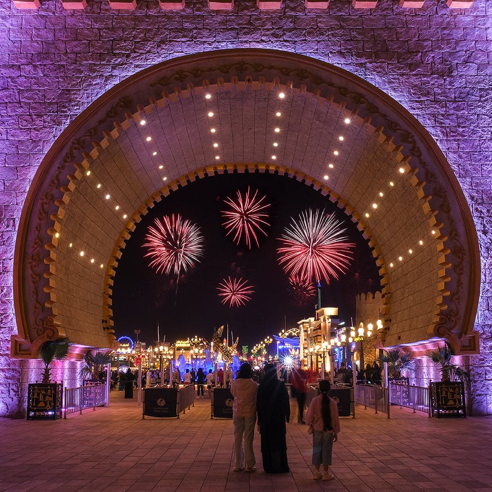 조명이 켜진 아치형 통로 아래에 서서 밤에 활기차고 조명이 밝은 놀이공원에서 활기찬 불꽃놀이를 감상하는 사람들.