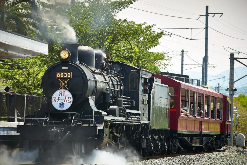 화창한 날 빨간 승용차를 끌어당기는 8630이라는 숫자를 가진 검은색 증기 기관차로, 나무를 배경으로 하고 있습니다.