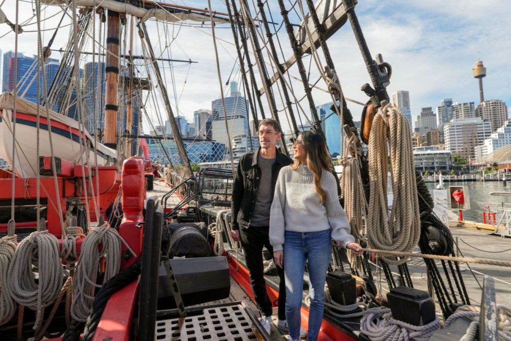 화창한 날 도시의 스카이라인을 배경으로 유서 깊은 배의 갑판을 걷고 있는 두 사람.