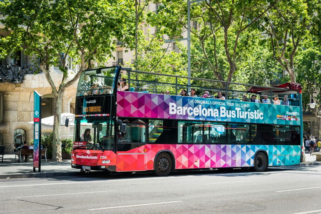 "Barcelona Bus Turistic" 이라고 표시된 다채로운 2층 버스가 상부 갑판에 있으며, 나무가 늘어선 거리에 주차되어 있습니다.
