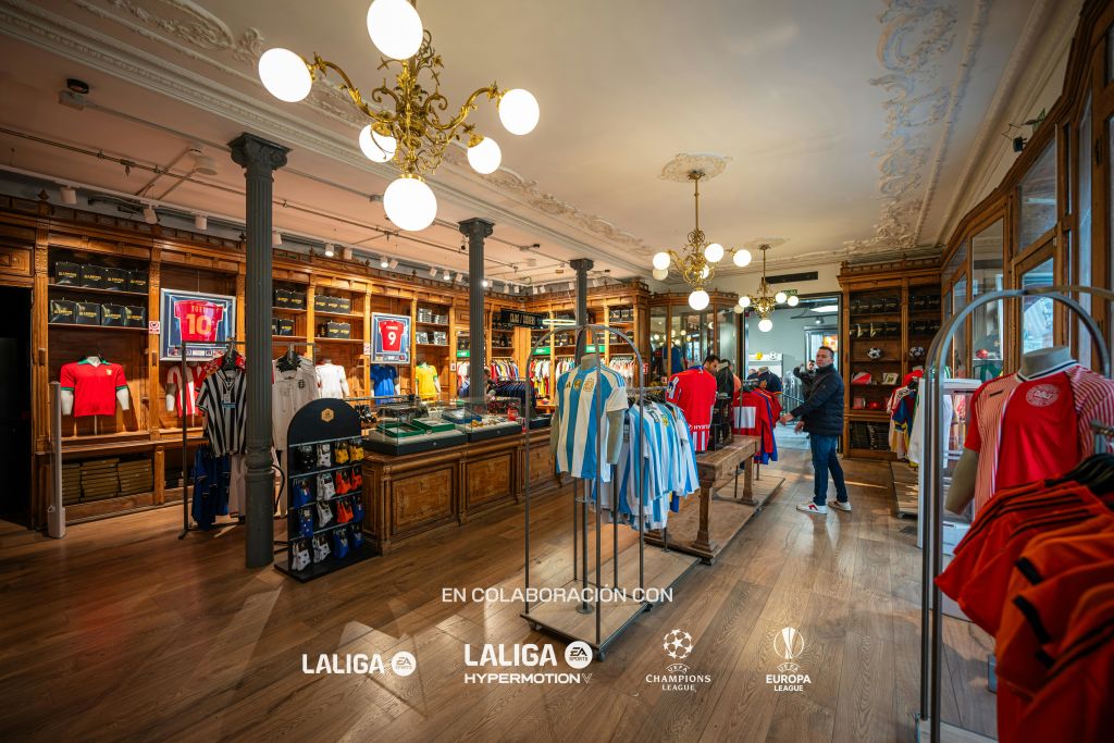 다양한 유니폼이 진열된 스포츠 용품점, 화려한 조명기구, 쇼핑객 몇 명이 물건을 둘러보고 있습니다.