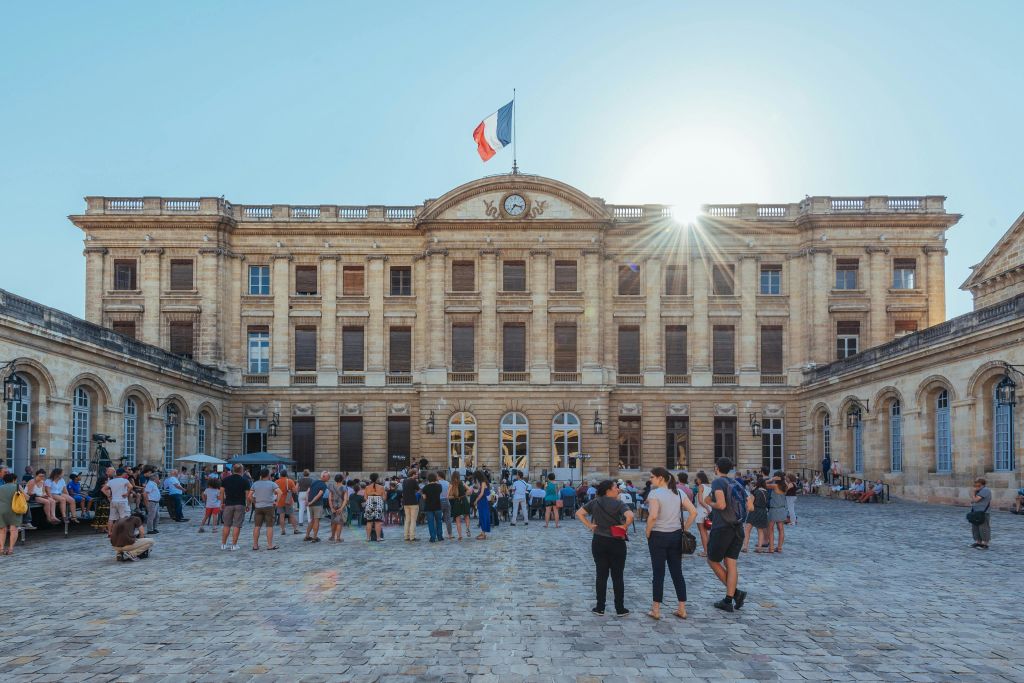건물 위에는 프랑스 국기가 있고, 햇살이 내리쬐는 역사적인 건물이며, 안뜰에는 많은 사람들이 모여 있습니다.