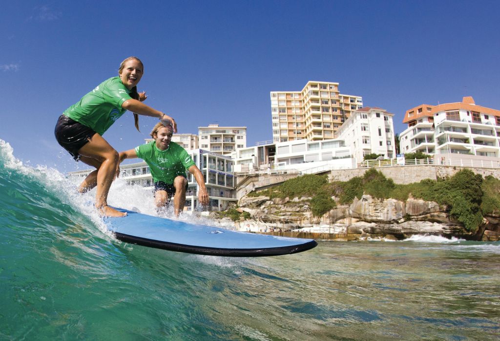 녹색 셔츠를 입은 두 사람이 건물을 배경으로 해안가 근처의 파도에서 파란색 서핑 보드를 타고 있습니다.