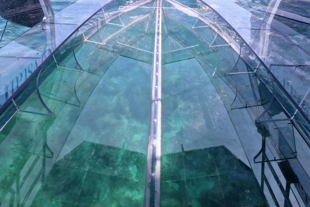 선박의 투명한 부분을 통해 바다가 보이는 산호초 위를 항해하는 투명한 바닥의 보트입니다.