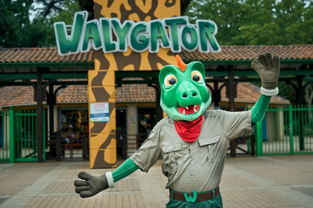 드래곤 코스튬을 입은 사람이 "WALYGATOR" 라고 적힌 표지판이 있는 놀이공원 입구 앞에서 손을 흔들고 있습니다.